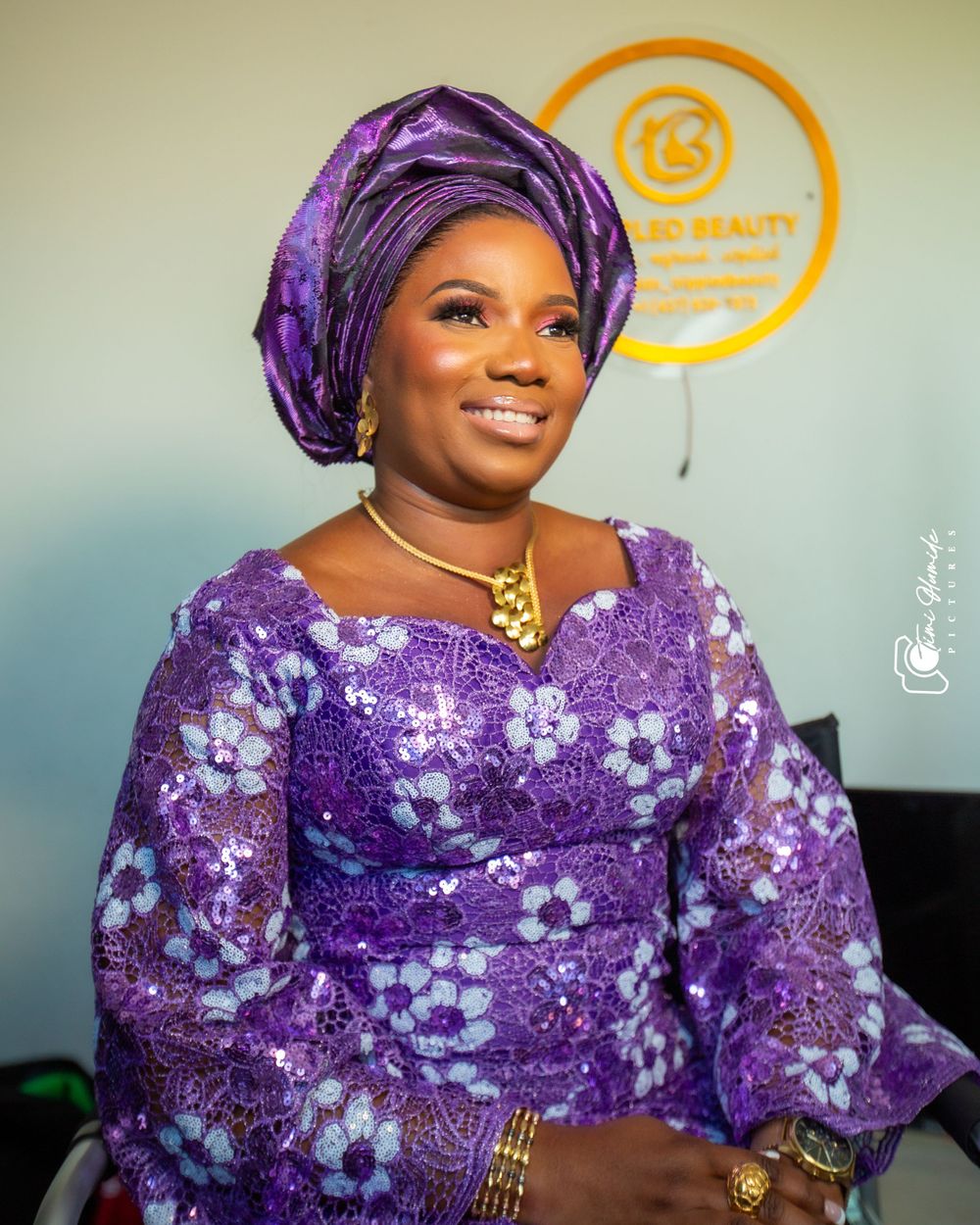 A woman in purple traditional African attire poses in front of a circular orange logo.