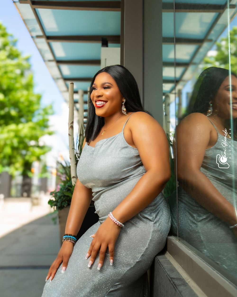 A woman in a silver dress poses against a reflective glass window outdoors.