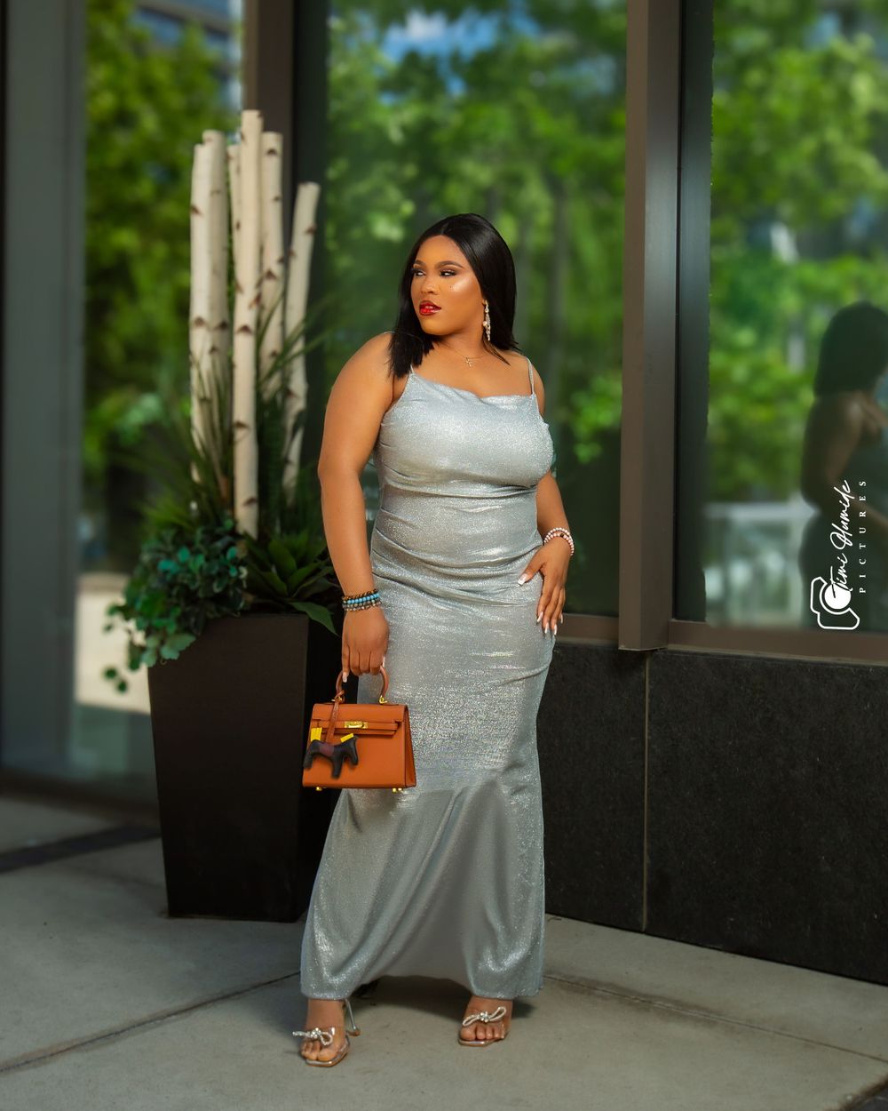 A woman in a fitted silver gown poses with a brown purse near modern architecture.