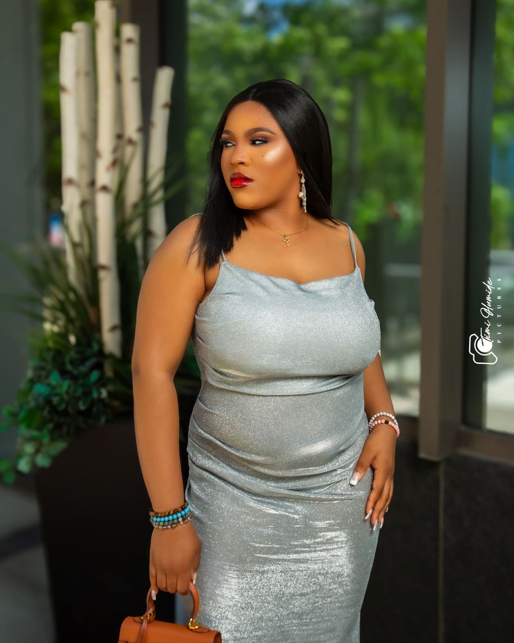 A woman in a silver sparkly dress poses with a brown handbag in an elegant setting.