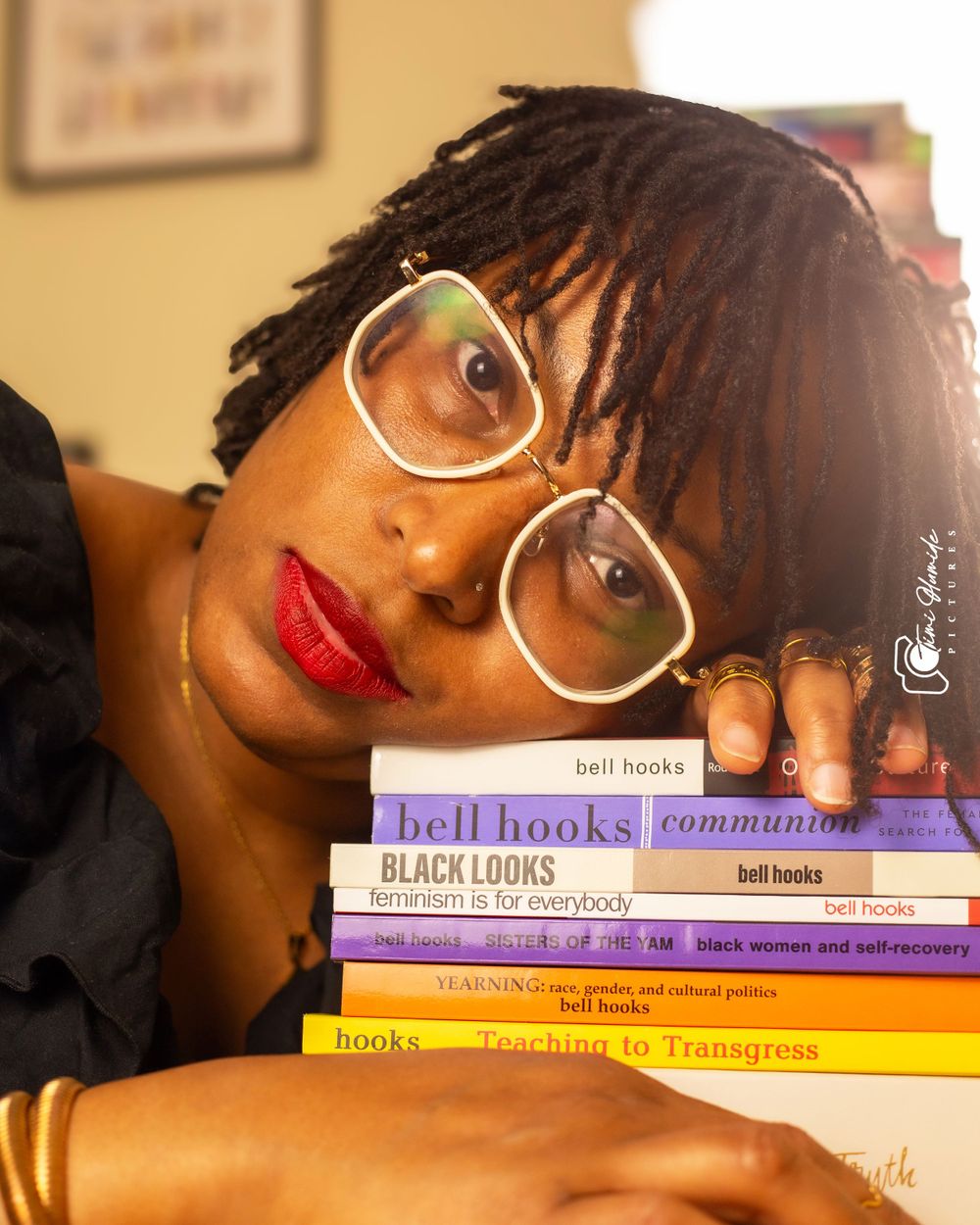 A person rests their head on a stack of bell hooks books while wearing white-rimmed glasses.