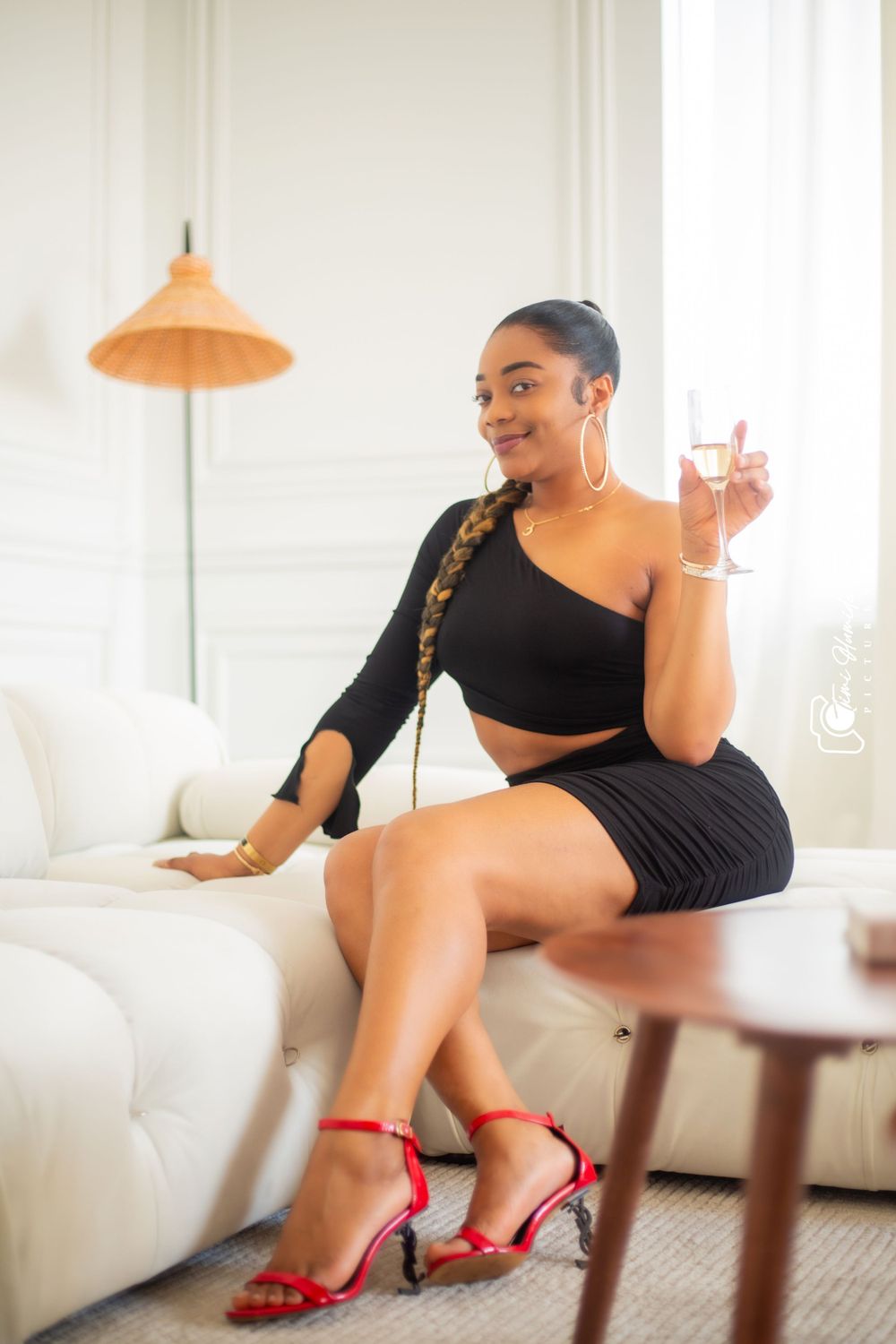 Woman in black crop top and skirt sits on white couch with champagne glass.