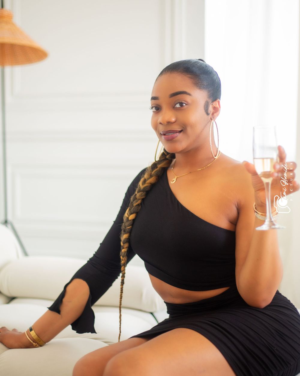 Woman in black crop top ensemble poses with champagne on white couch.