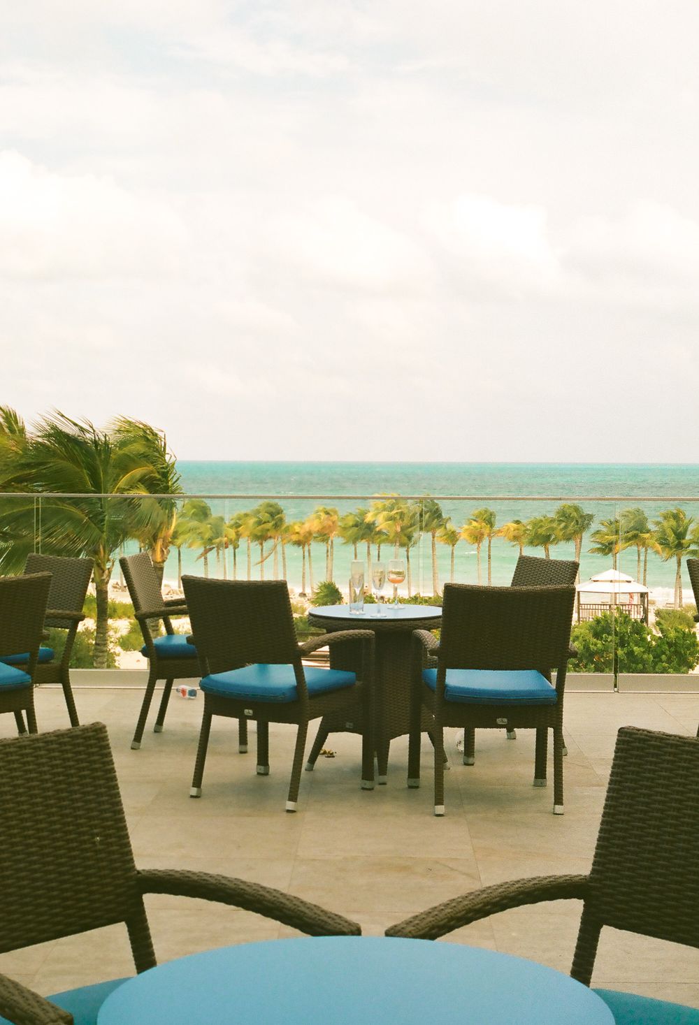 Outdoor resort dining area with wicker furniture overlooking palm trees and beach.