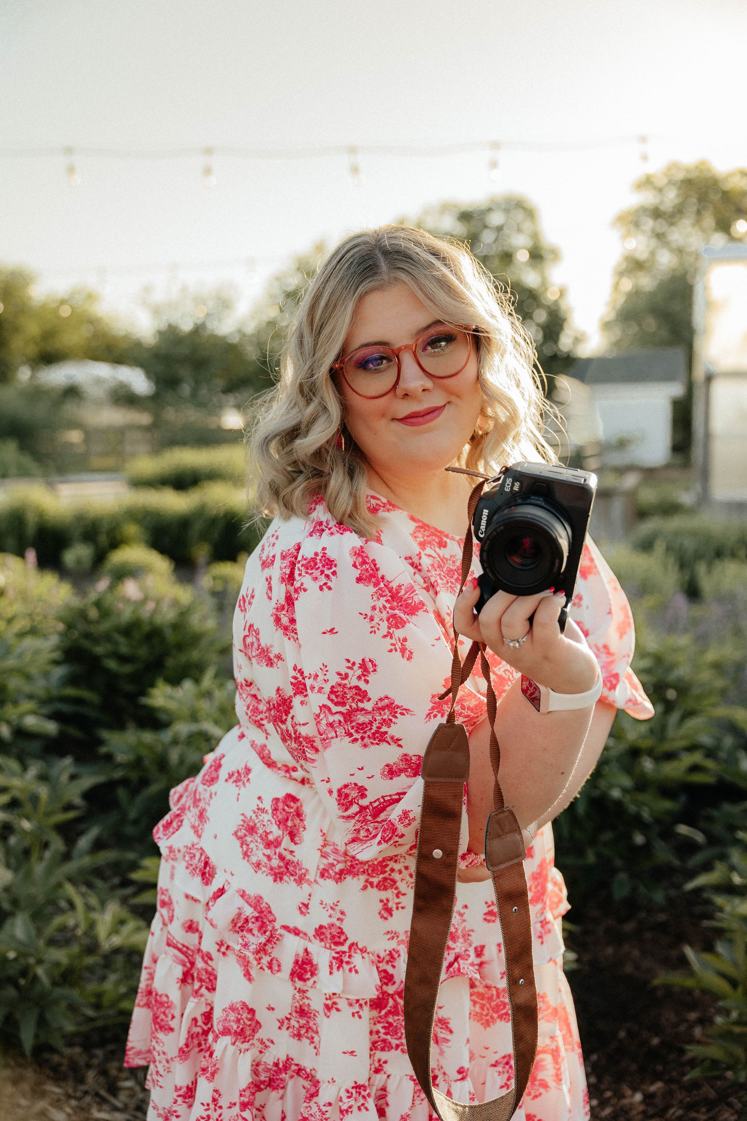 Blonde woman in pink floral dress holding camera outdoors in garden at golden hour.