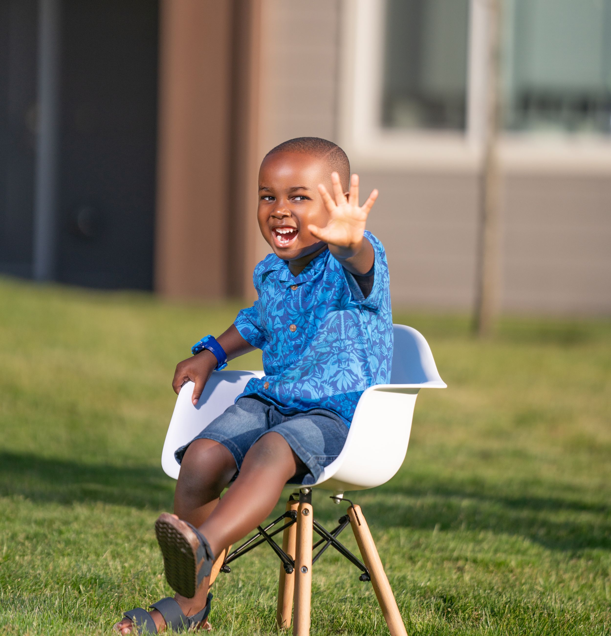 A young child in a blue shirt waves happily while sitting on a modern white chair outdoors on grass.