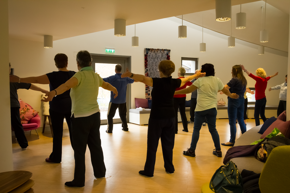 Group of people stretching with arms outstretched in a dance studio with mirrors and pendant lights.