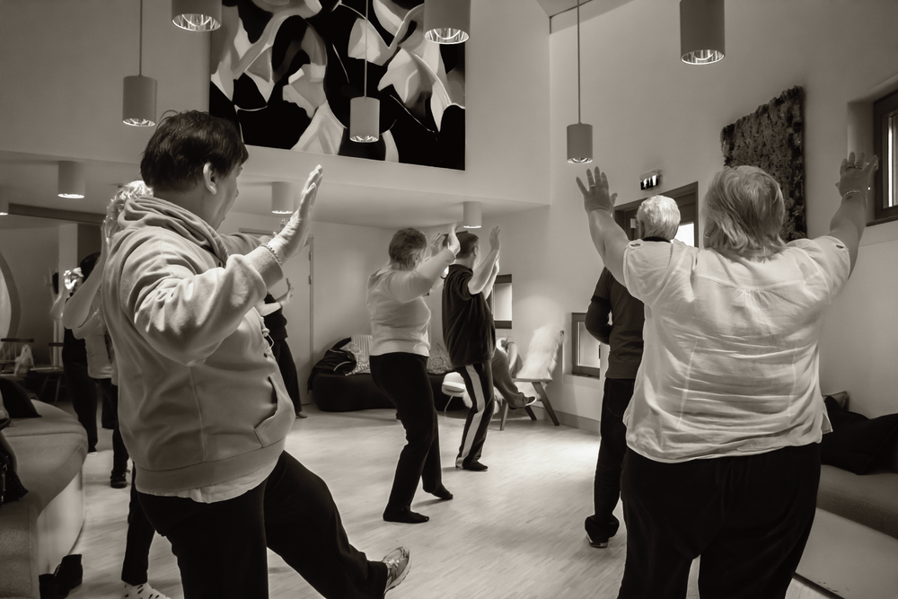 People exercising in a dance studio with mirrors and overhead lighting in black and white.