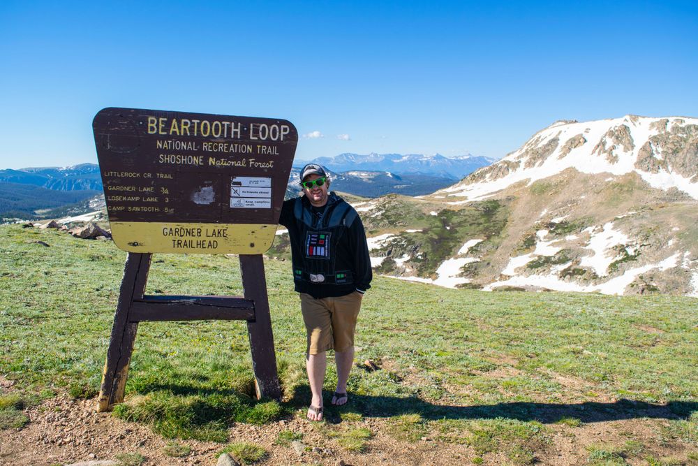 Wooden Beartooth Loop informational sign on mountain summit with snowy peaks in background.