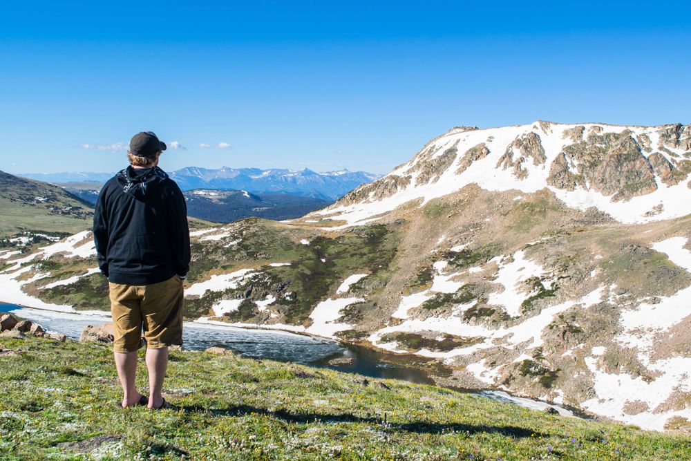 A hiker stands on an alpine ridge overlooking snow-patched mountains and valleys under a bright blue sky.