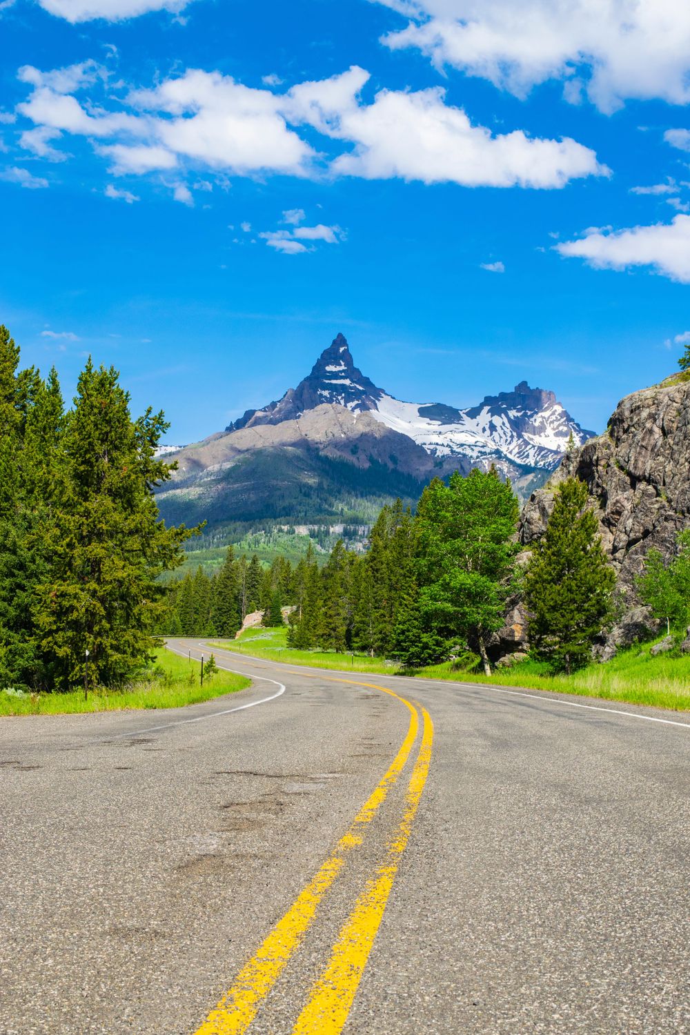 A winding mountain road leads toward a dramatic peaked mountain under blue summer skies.
