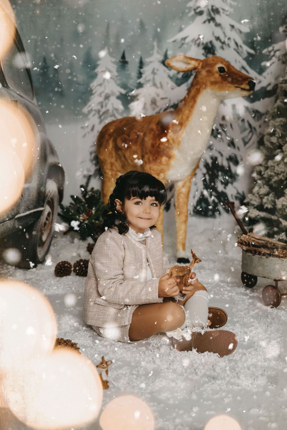 A whimsical winter portrait in a snowy forest scene with a deer decoration and bokeh lighting effects.