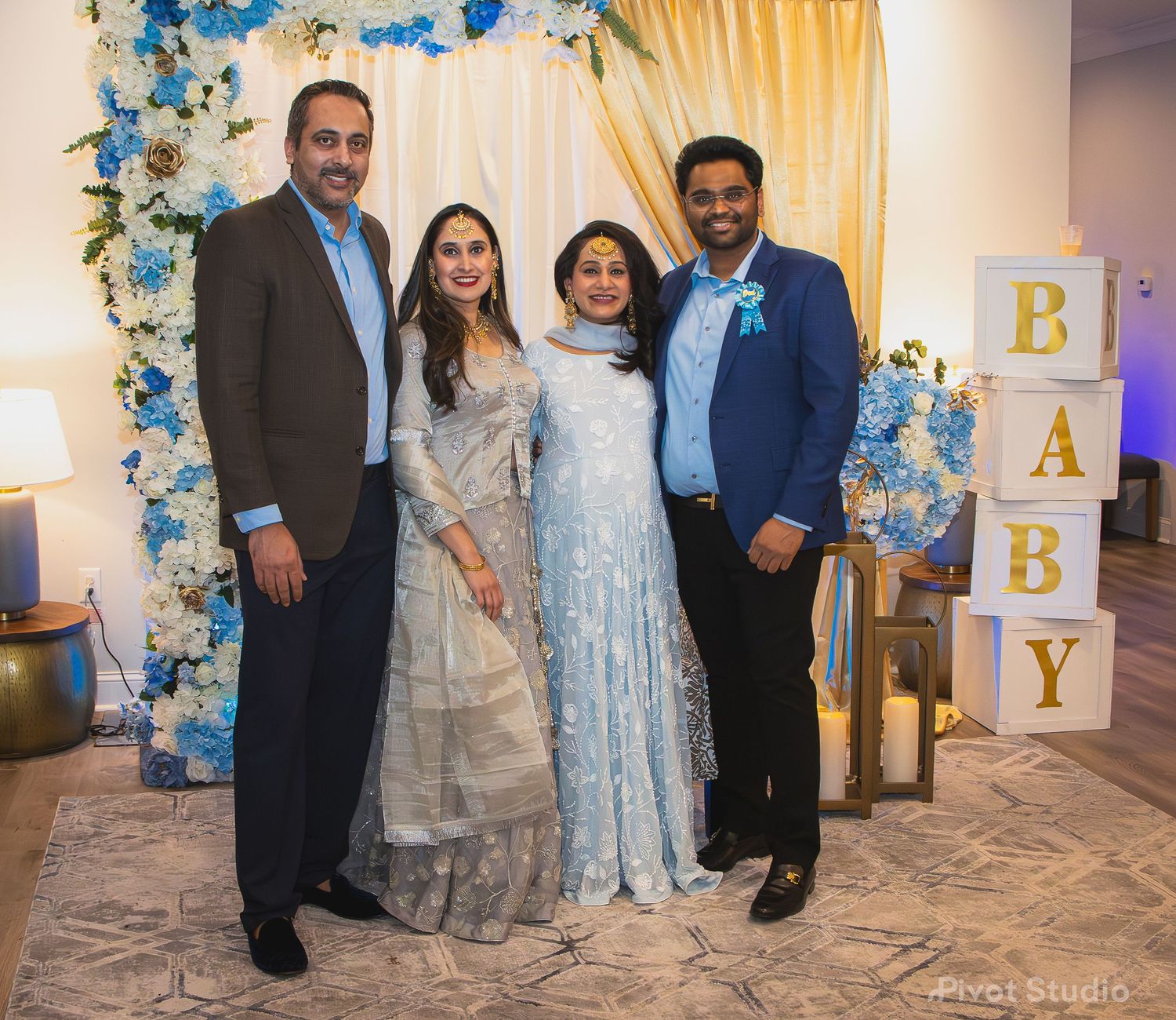 Four people posing together at a baby shower event with blue and white floral decorations and BABY blocks.