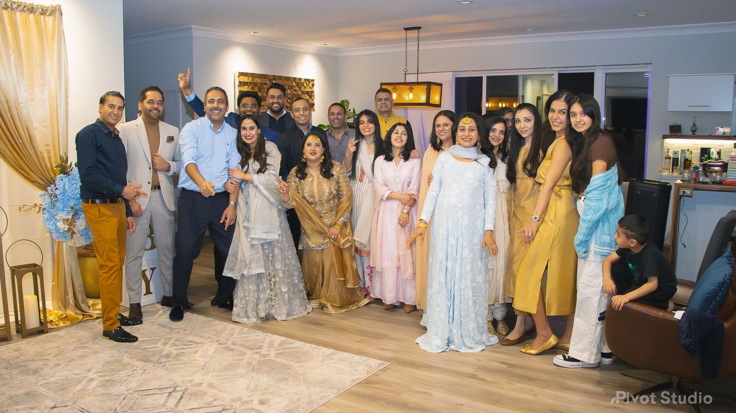 Large South Asian family group photo at an elegant indoor celebration with gold decor.