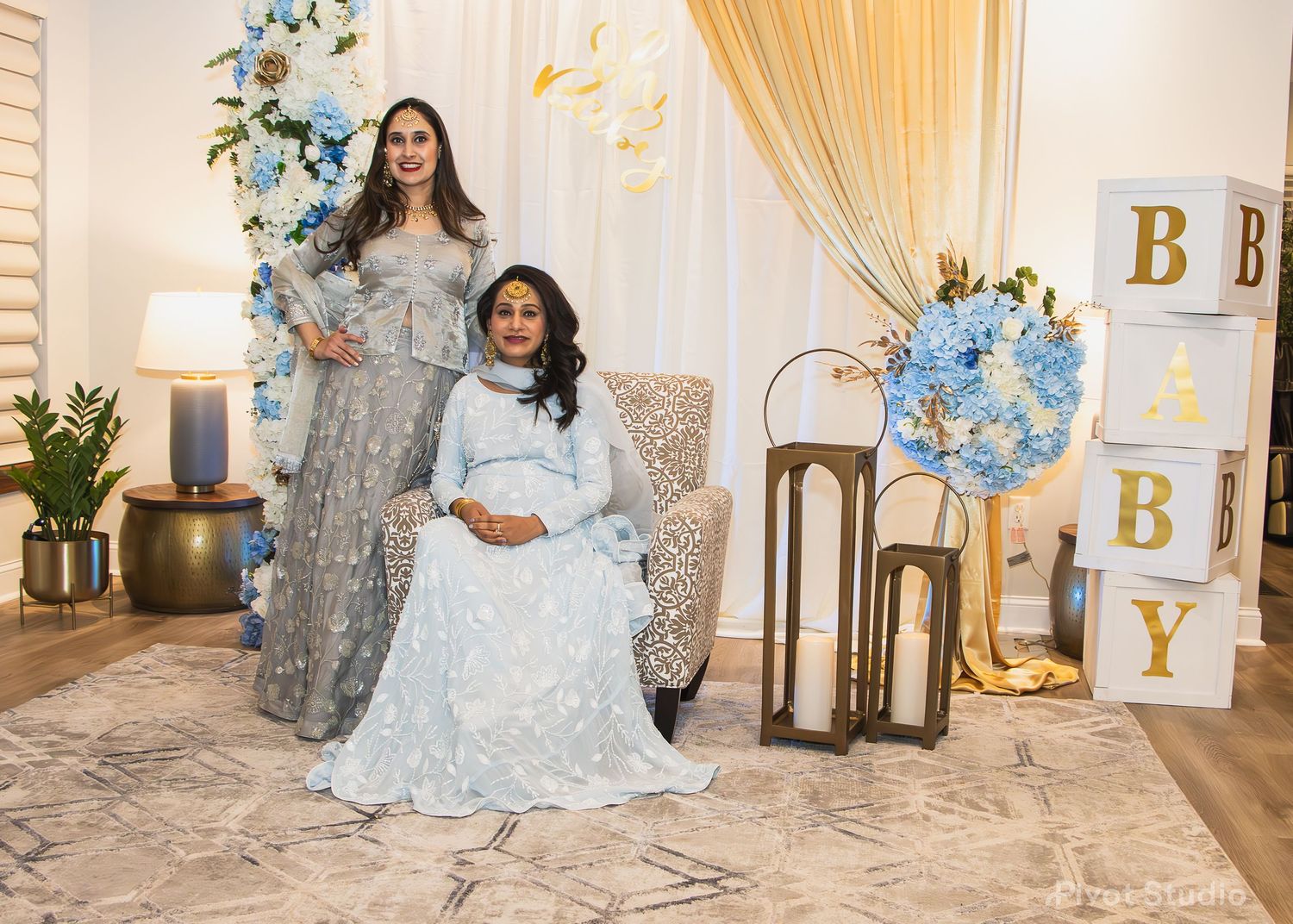 Two women in elegant gray dresses pose at a blue and gold baby shower decoration setup.