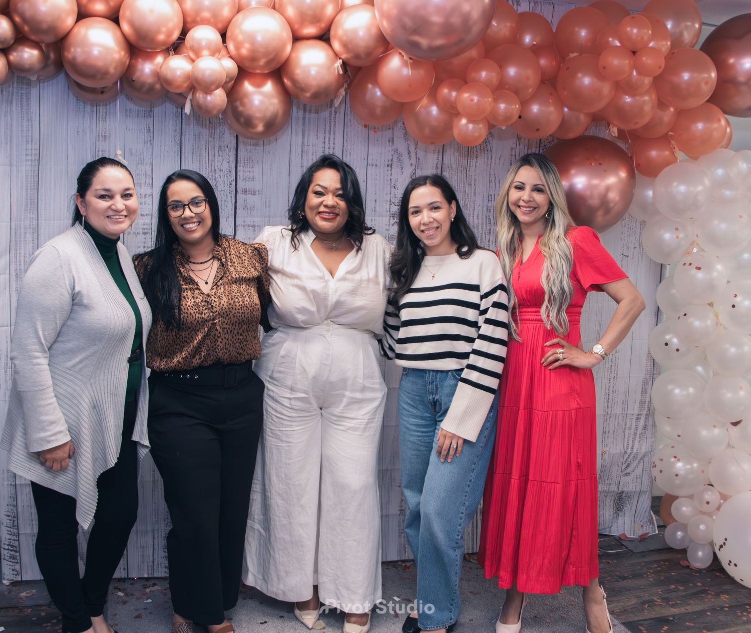 Five women smiling together under a rose gold balloon arch at a celebration event.