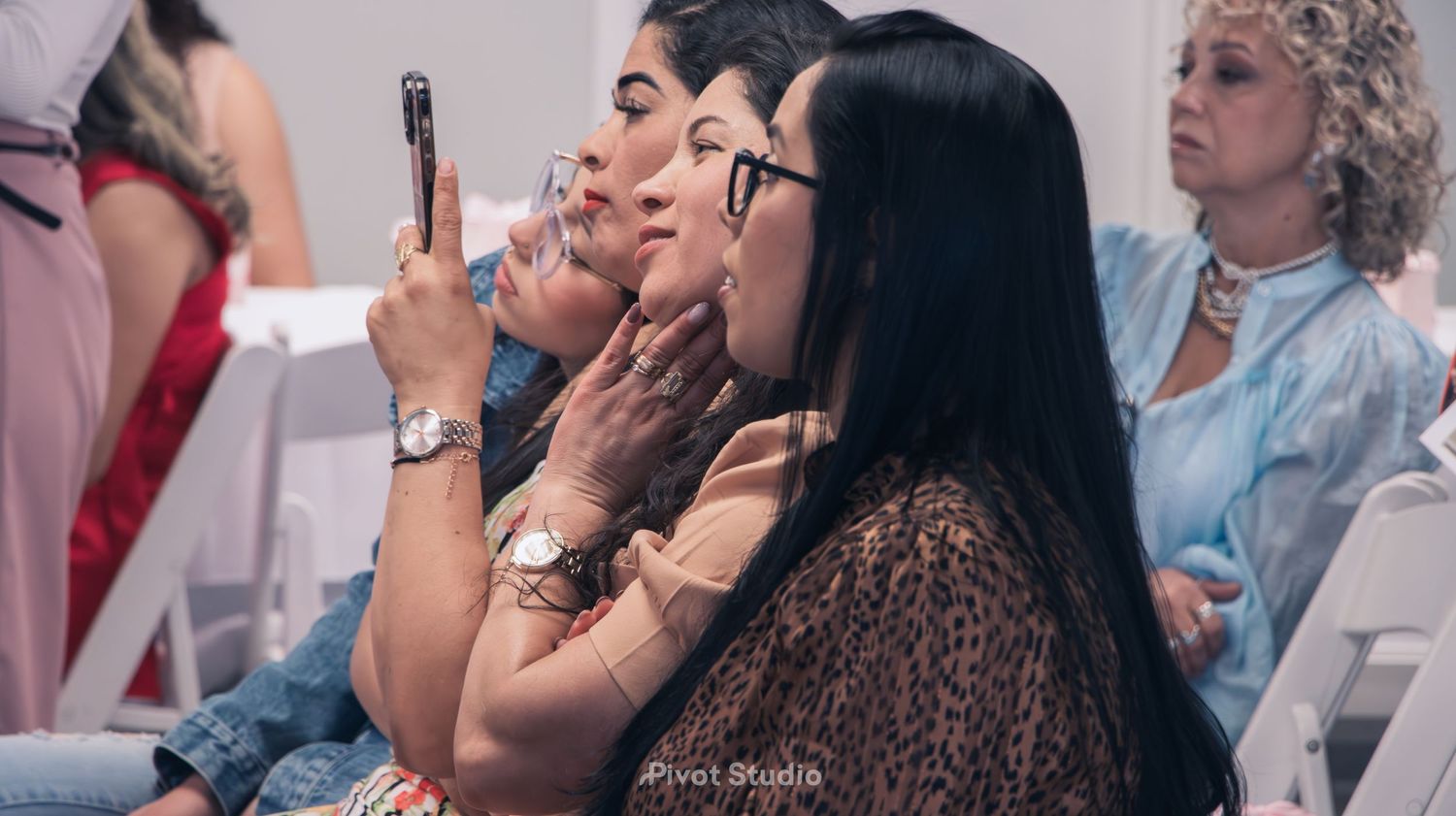 Women seated at an event, one recording on smartphone, wearing elegant patterned outfits.