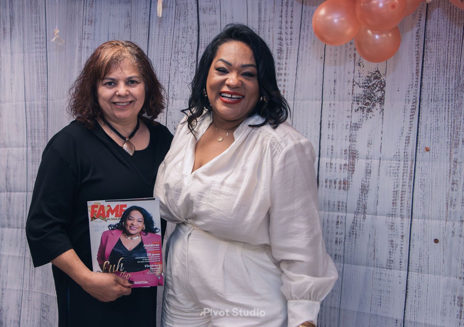 Two women pose together holding a BAME magazine at a rose gold balloon decorated event.