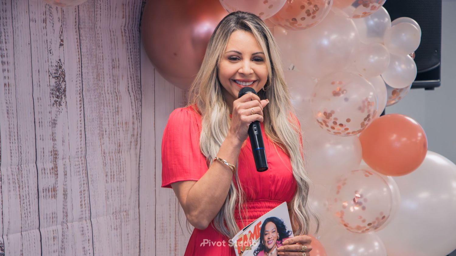 Blonde woman in red dress speaks into microphone holding magazine at balloon-decorated event.