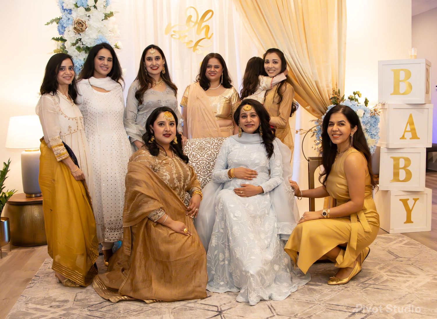 Group of eight women in traditional South Asian attire posing at a baby shower event.