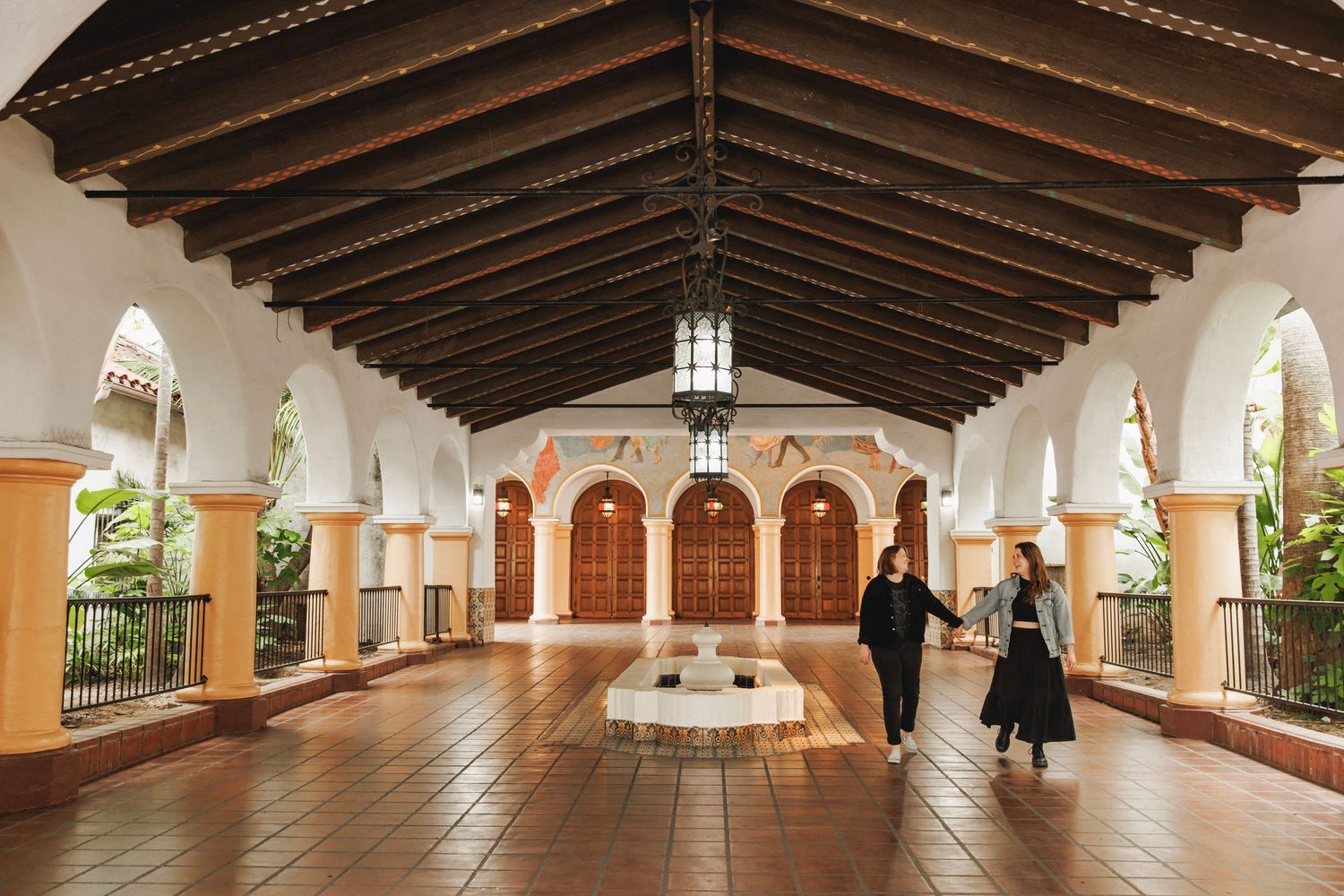 Couple walking through a grand Spanish colonial hall with arched ceilings and tile floors.