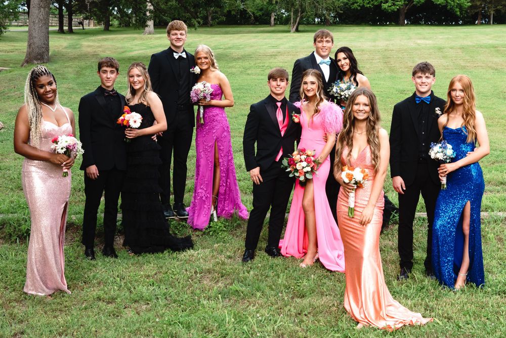 Large group in formal attire poses for prom photos on grass lawn.