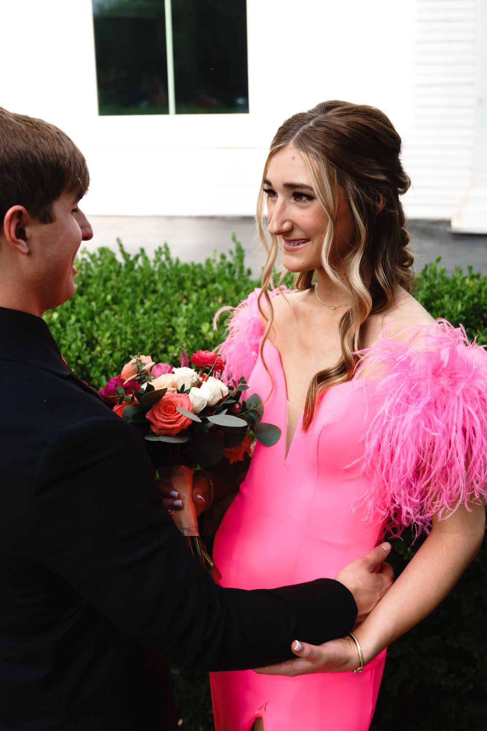 Young couple in formal attire with woman wearing vibrant pink feathered dress holding flowers.
