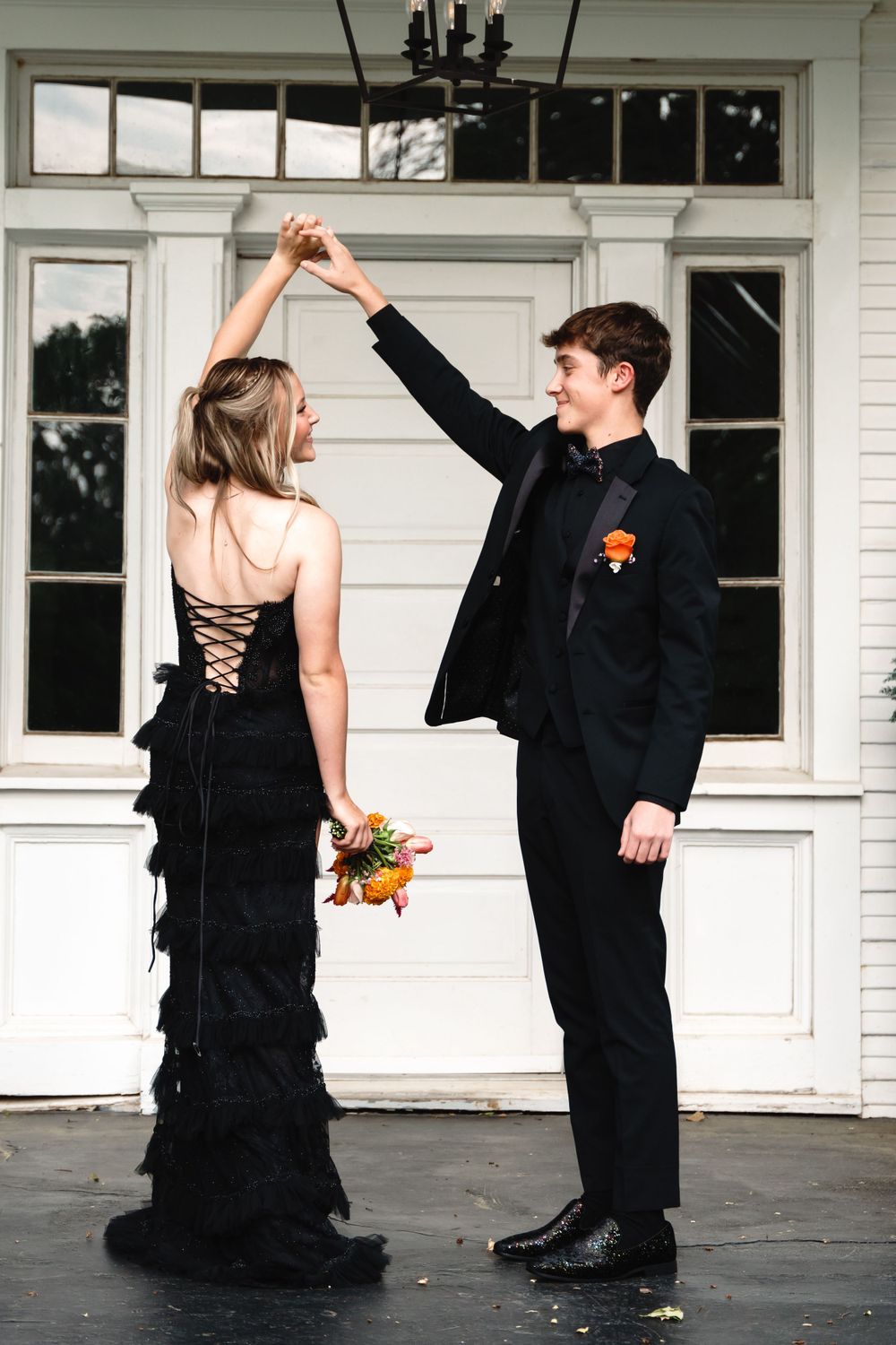Couple in black formal wear performs dance twirl on white porch.