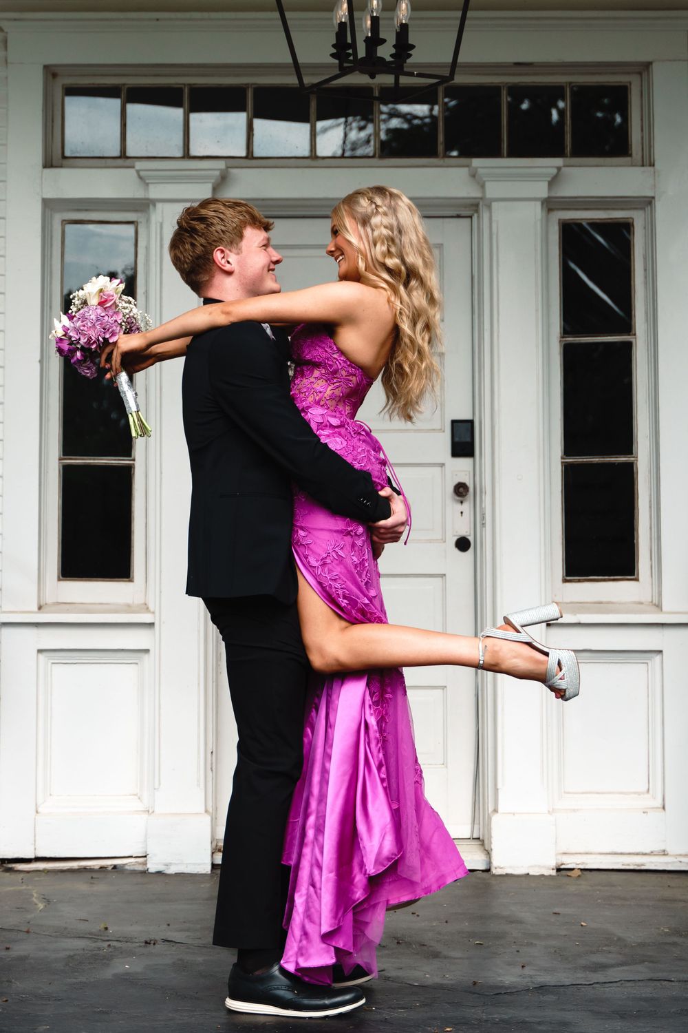 Playful prom couple embracing in front of white doors, with the woman wearing a pink sequined dress.