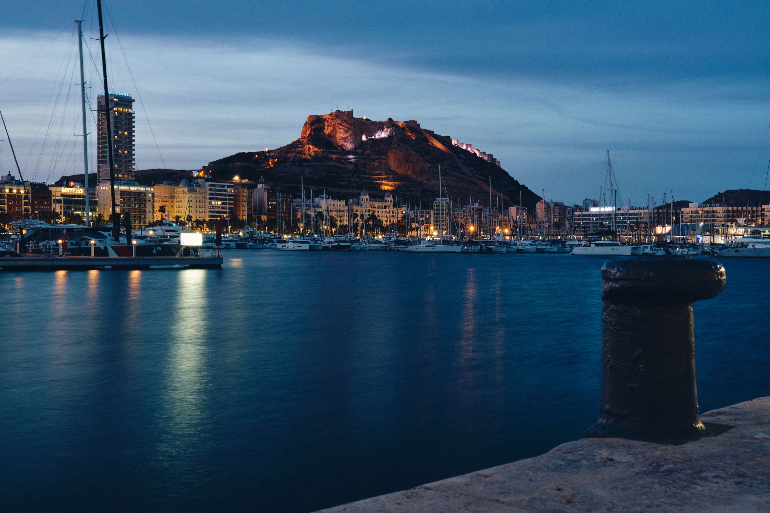 Night view of illuminated marina and castle in Alicante with boats reflecting on calm waters.