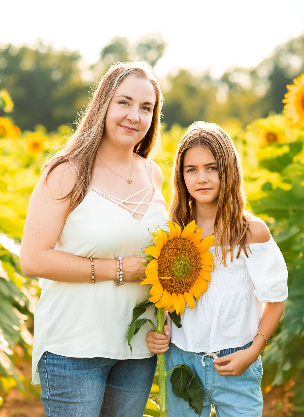 Mother and daughter in matching denim and white tops pose together in a blooming sunflower field.