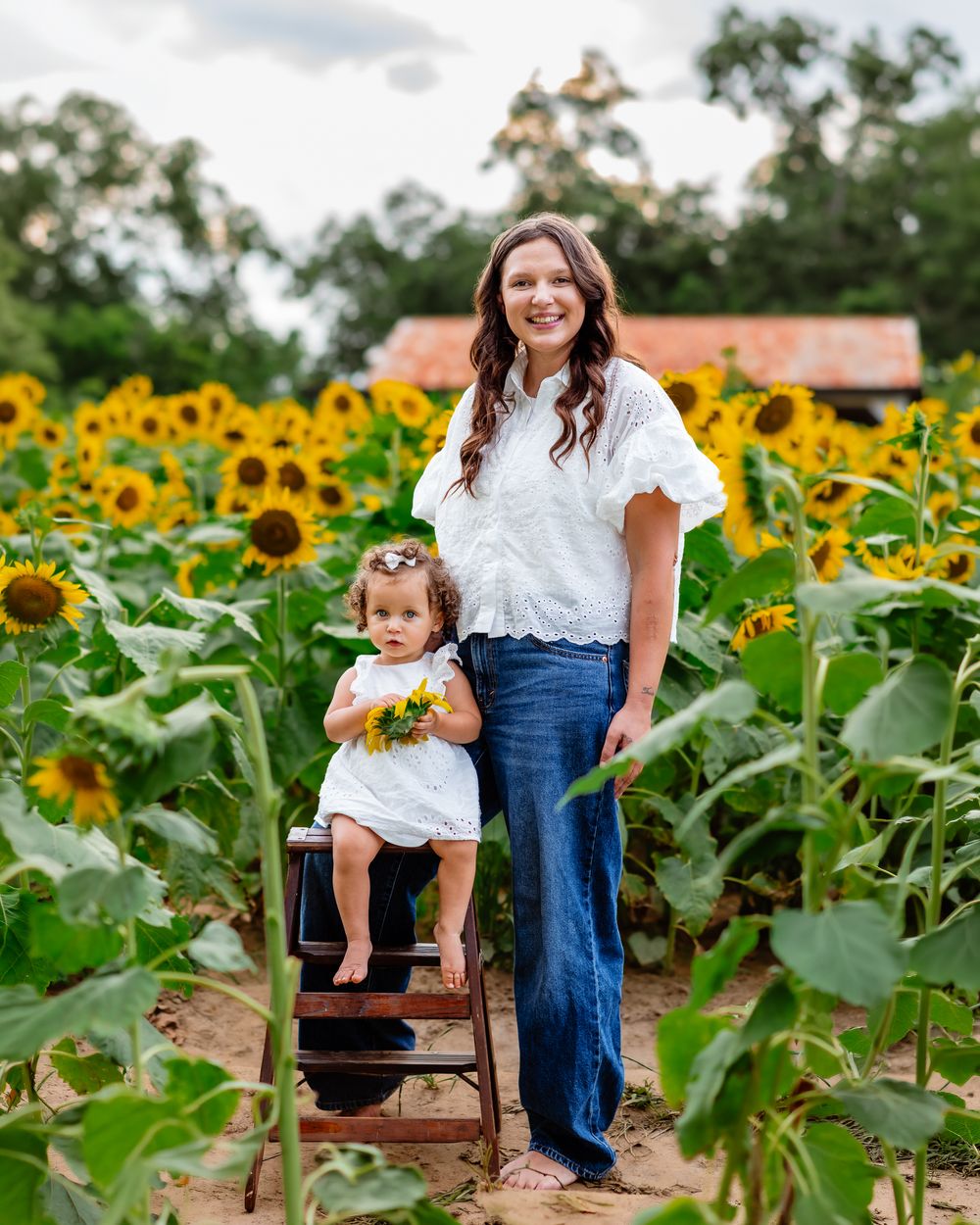 Woman and toddler in matching white outfits posing in a vibrant sunflower field, child holding yellow flowers.
