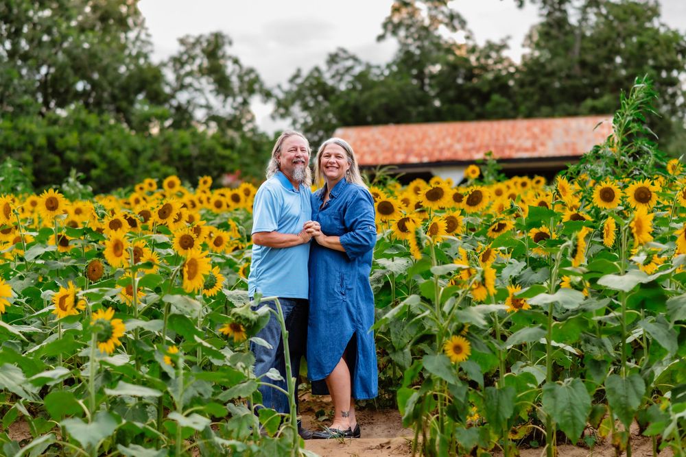 Couple smiling together in a vibrant sunflower field, wearing blue outfits, with a rustic barn in the background.