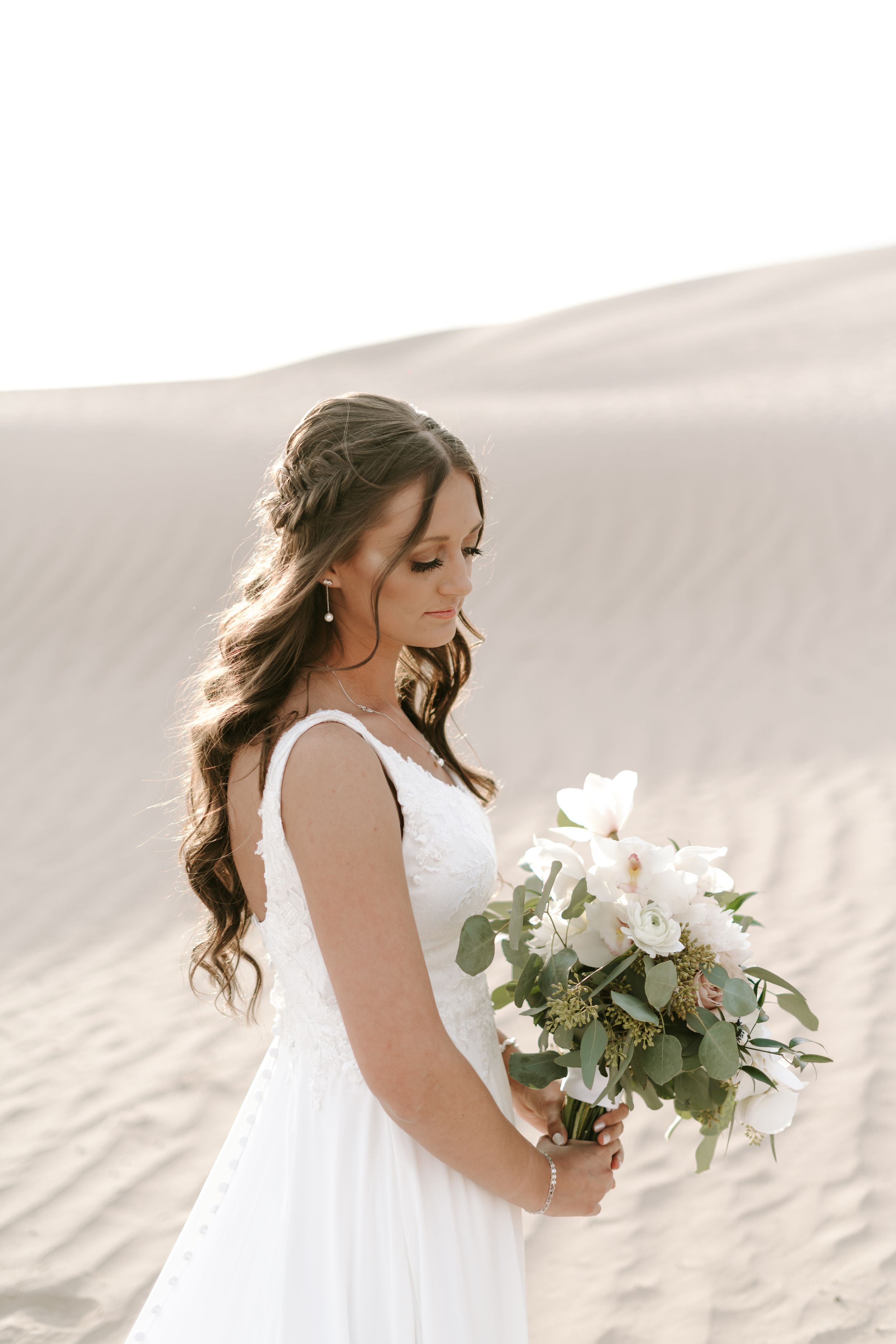 Bride in flowing white dress holds elegant white bouquet against sand dune backdrop.