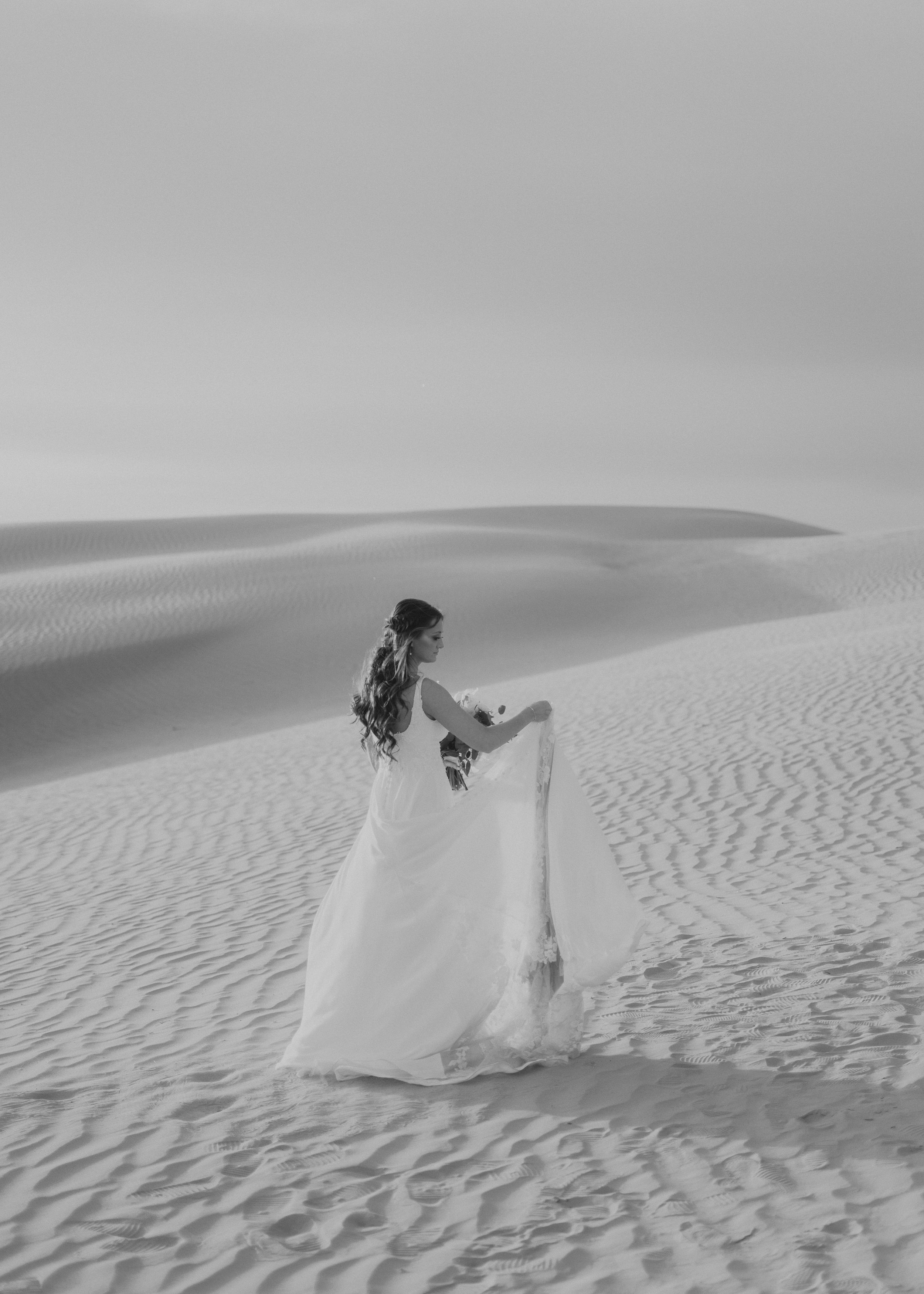Black and white photo of bride in flowing dress standing alone on desert sand dunes.