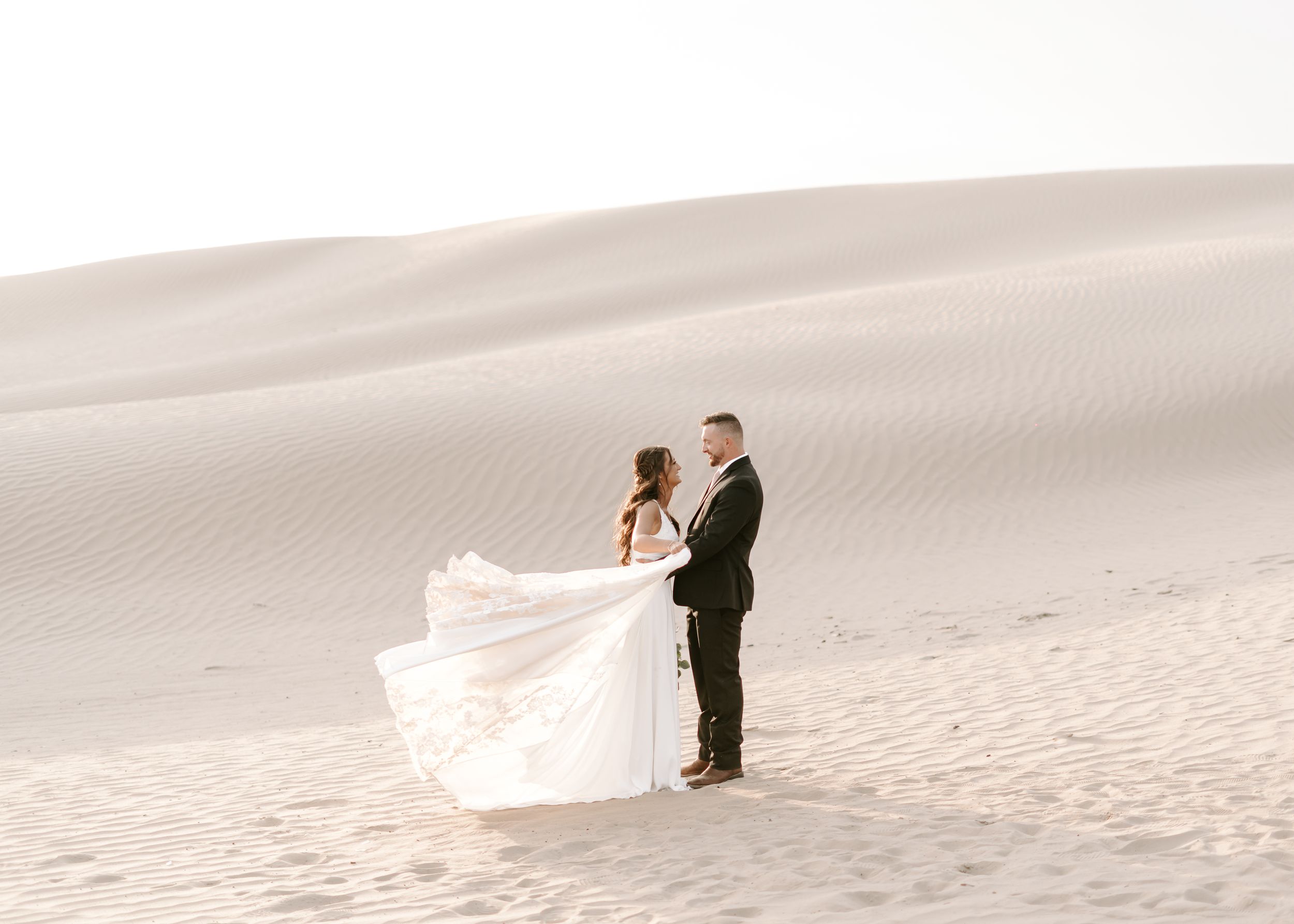 Wedding couple stands together on sandy dunes as bride's dress flows in the wind.