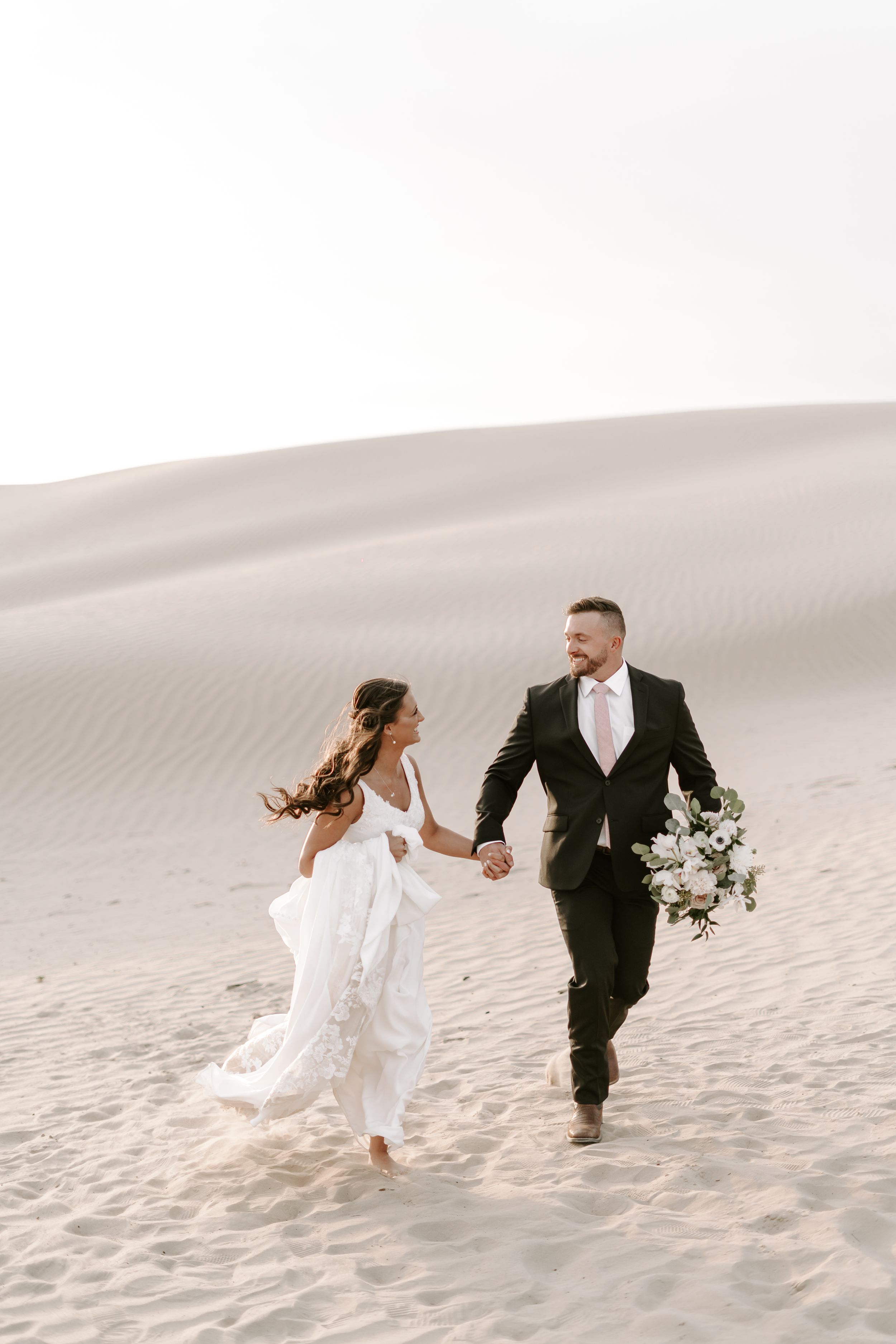 Newlyweds run hand in hand across desert sand dunes in wedding attire.