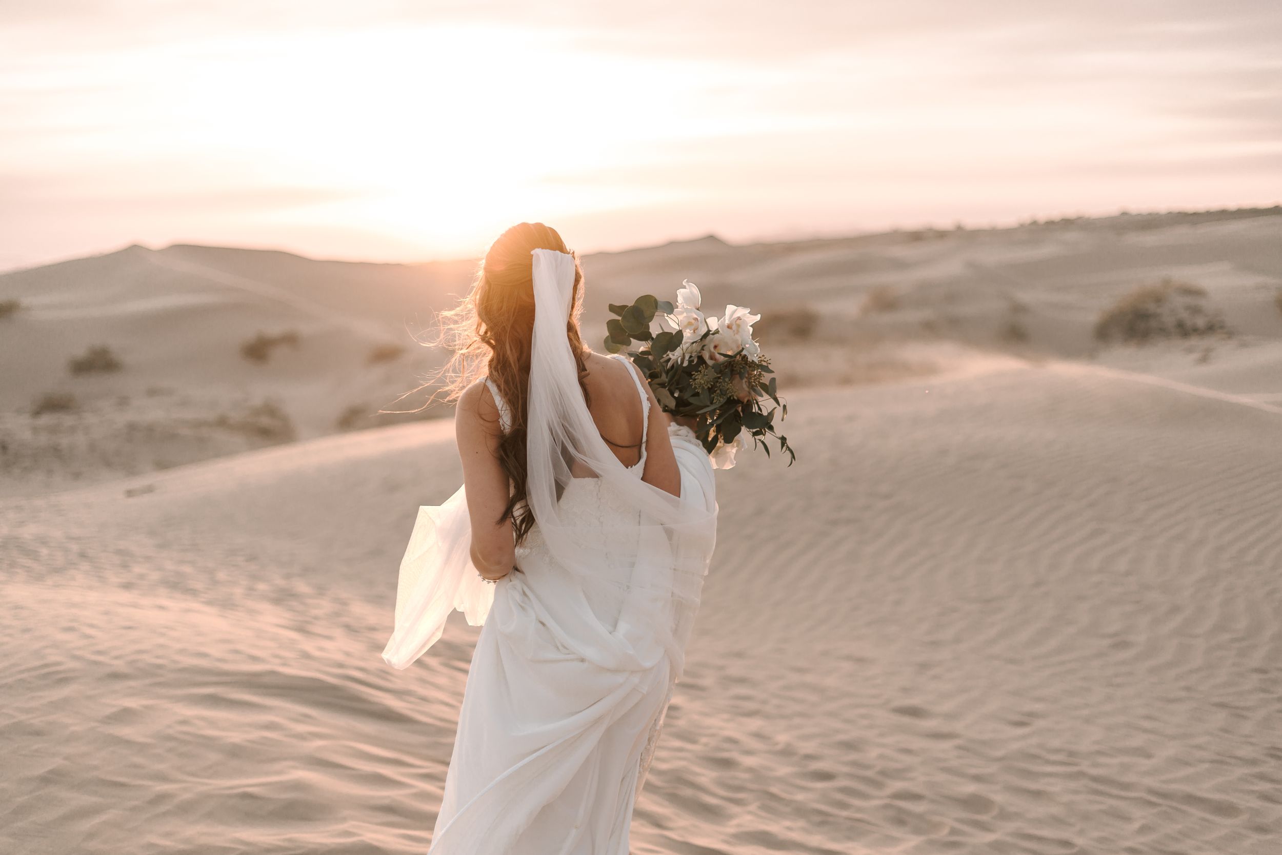 Elegant bride in white dress holding bouquet against sunset desert landscape.