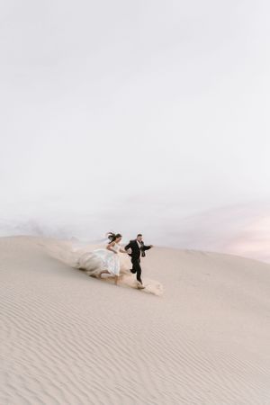 Joyful newlyweds running together across desert sand dunes at sunset.