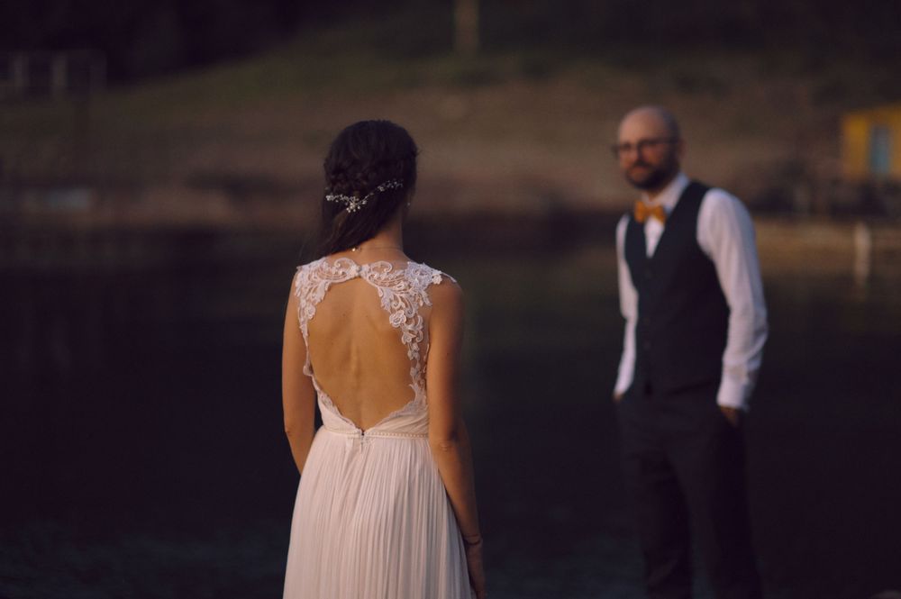 Back view of a bride in an open-back lace wedding dress standing with her partner near the water.