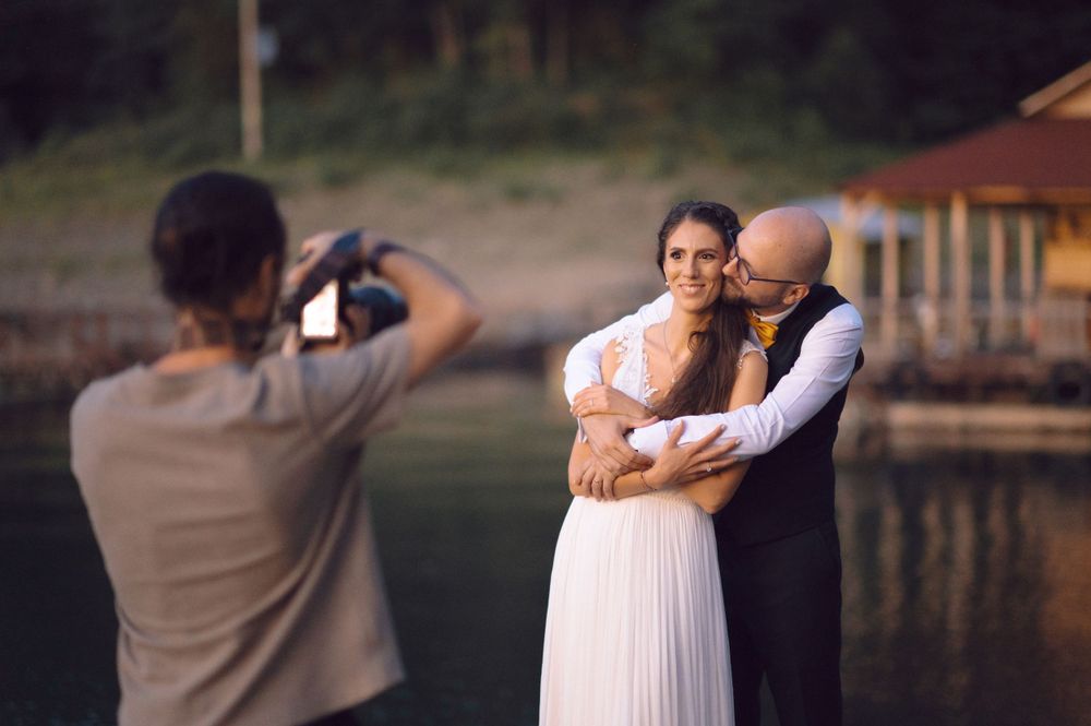 Behind the scenes of a wedding photoshoot by a lake with photographer capturing the couple.