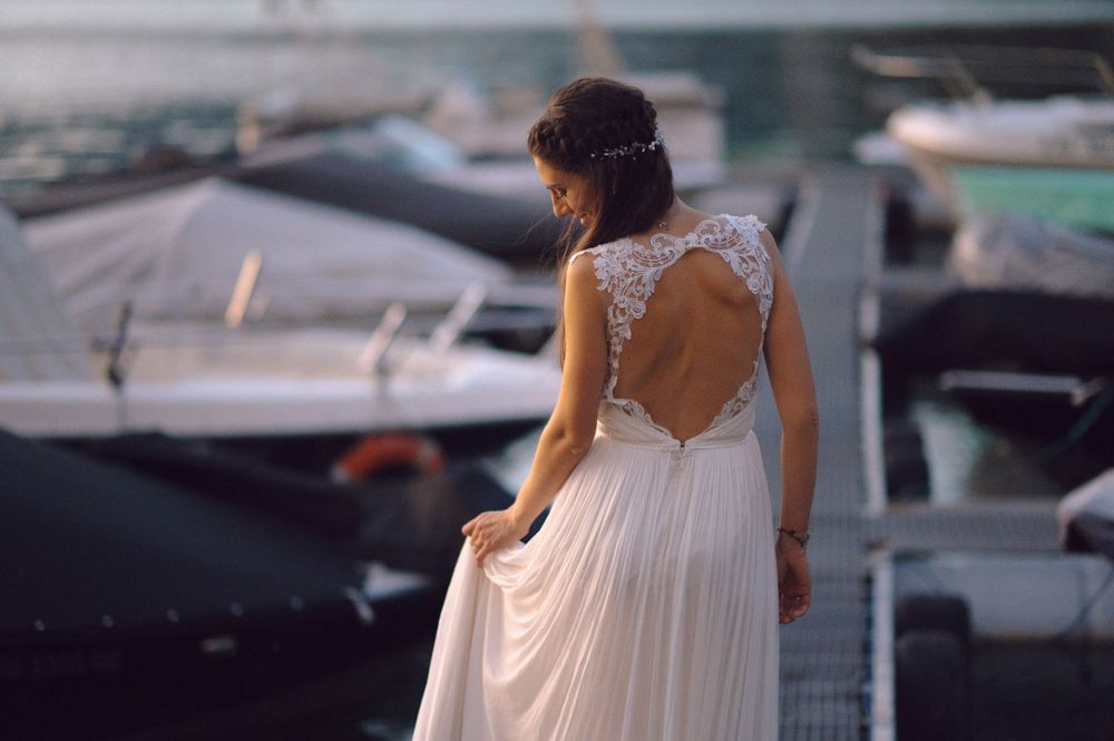 Bride in a flowing backless wedding dress standing near boats at a marina.
