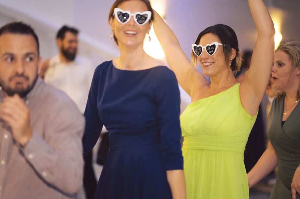 Women wearing sunglasses and colorful dresses dancing enthusiastically at party.