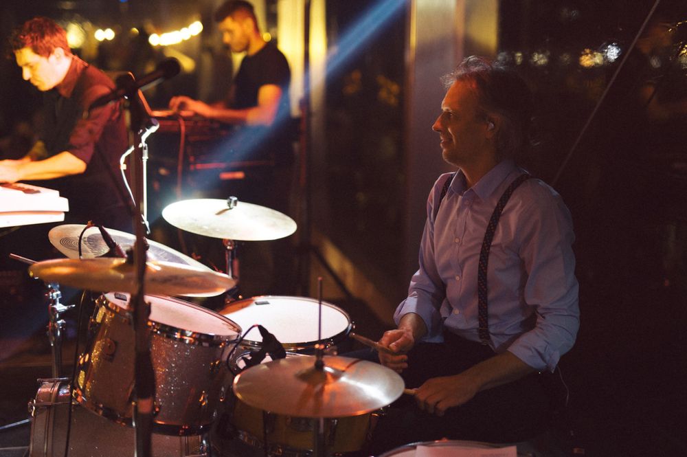 A drummer performs behind a drum kit in moody stage lighting.