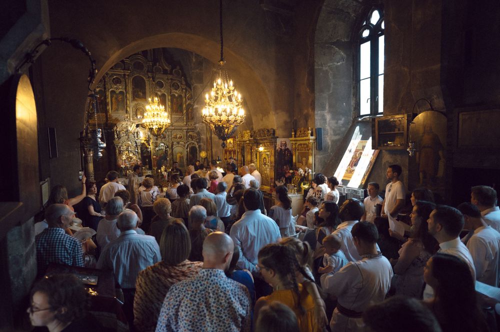 A crowded Orthodox Christian church service with chandeliers and religious icons visible in the dimly lit space.