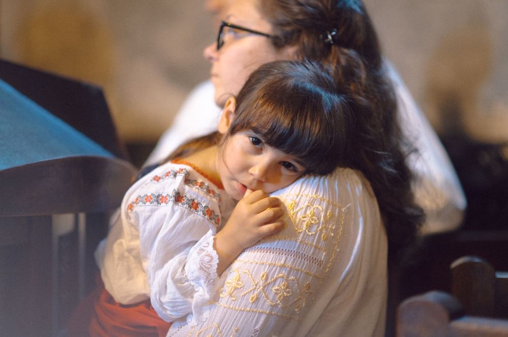 A child rests while wearing an embroidered white folk dress during a traditional event.