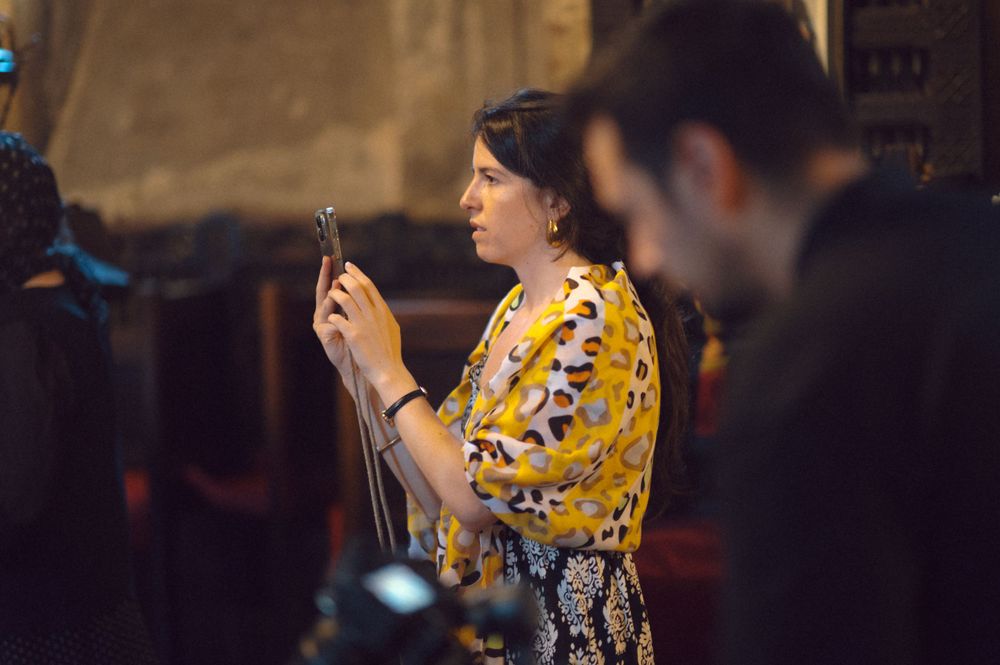 A person in a yellow patterned dress takes photos during an indoor event.