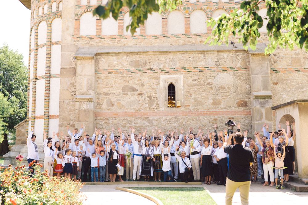 A large group wearing traditional Romanian clothing poses in front of a historic stone church.