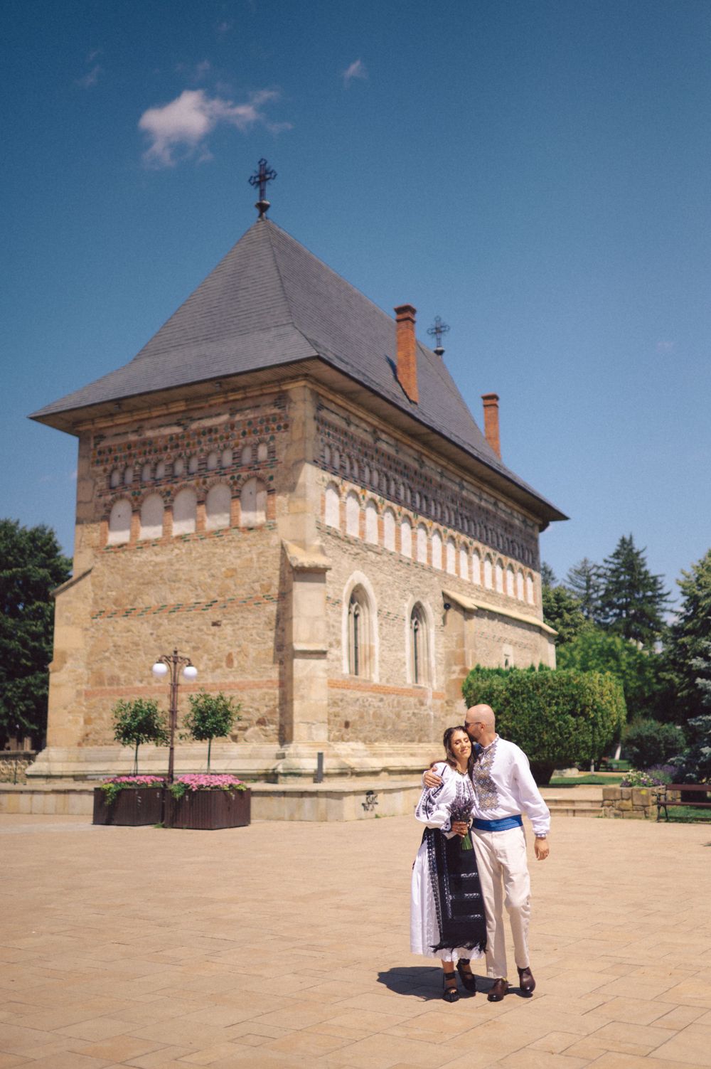 A couple stands in traditional attire in front of a historic Romanian Orthodox church.