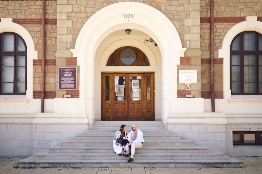 A couple in traditional Romanian dress sits on stone steps of a historic building.